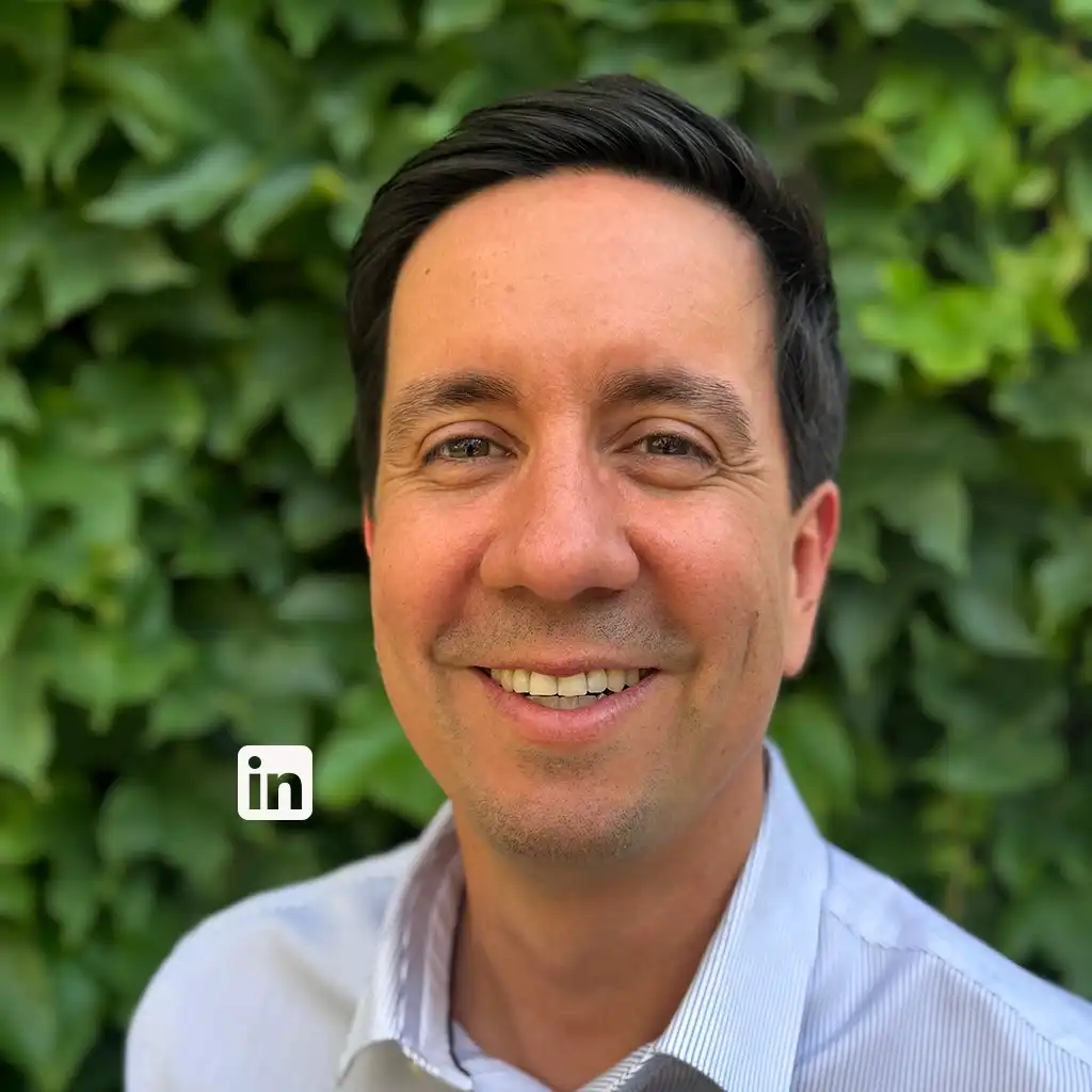 Smiling man in a light blue striped shirt, head-and-shoulders portrait outdoors with green leaves in the background; LinkedIn icon visible on the image.