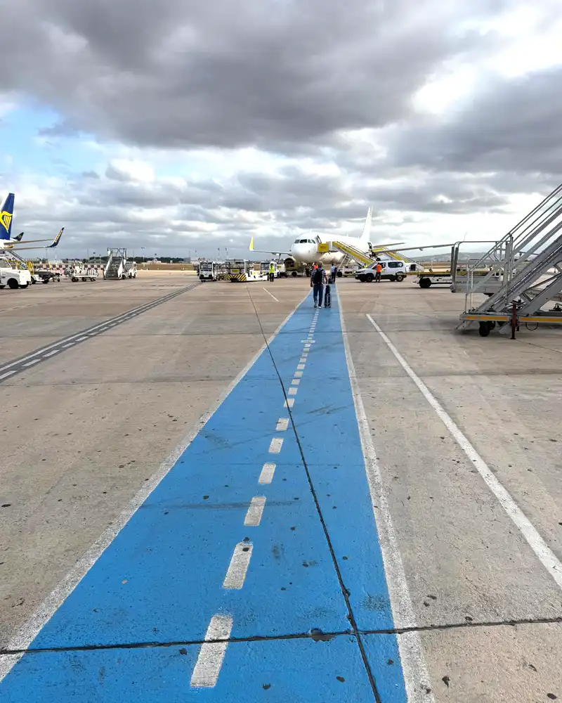 Airplane parked at a gate on the tarmac, a blue pedestrian path leads toward the jet bridge under cloudy skies.