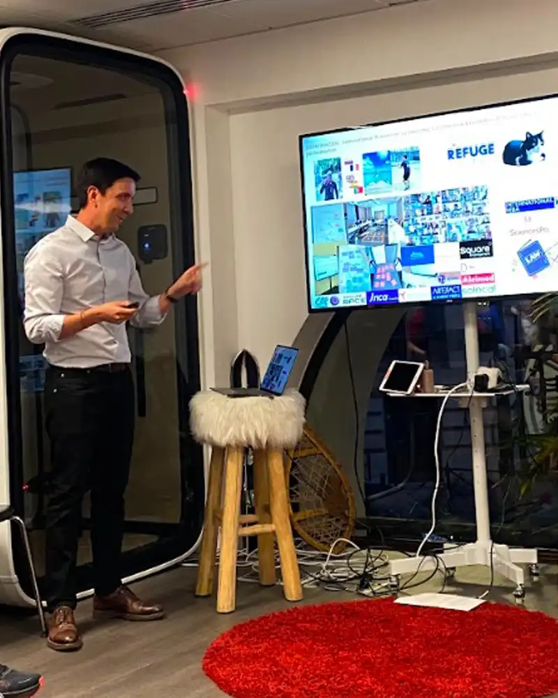 Man in a white shirt giving a presentation beside a large screen with a collage of slides in an office conference area.